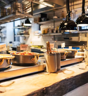 A modern restaurant kitchen with various pots of food, each labeled, lined up on a wooden counter. The background features white tiled walls, shelves with utensils and ingredients, and industrial-style lighting.