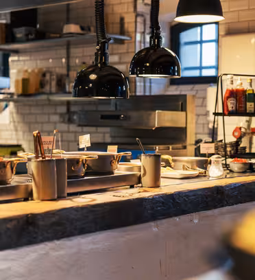 A cozy restaurant kitchen with a stone countertop displays various dishes in pots. The background features shelves with jars, condiments, a grill, and hanging lamps, under warm ambient lighting.