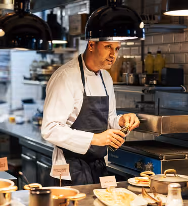 A chef in a white jacket and blue apron is seasoning food on a stovetop in a busy, well-lit kitchen. Plates of prepared dishes and condiments like ketchup and mustard are on the counter. Hanging lights illuminate the scene.