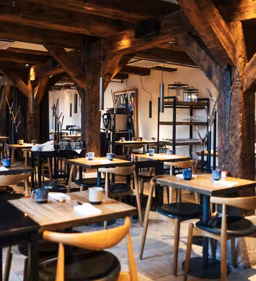 A cozy, rustic restaurant interior featuring wooden beams and tables with cushioned chairs. The tables are set with cups and napkins, creating an inviting atmosphere. Shelving with glassware lines the walls.