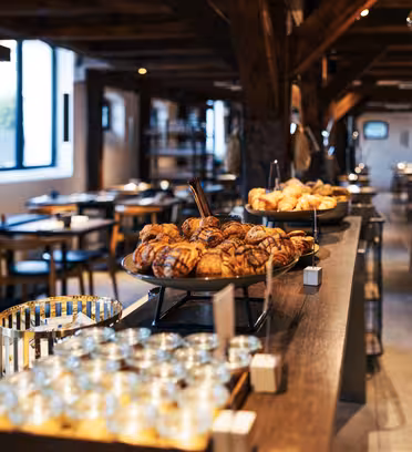 A cozy café interior featuring wooden beams and warm lighting. A central counter displays assorted pastries on trays, with small jars nearby. Wooden tables and chairs are arranged throughout the space, creating an inviting atmosphere.