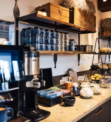 A cozy coffee station in a cafe features a coffee machine, stacked blue cups, and shelves with plants, snacks, and condiments. Wooden accents and soft lighting create a warm atmosphere.