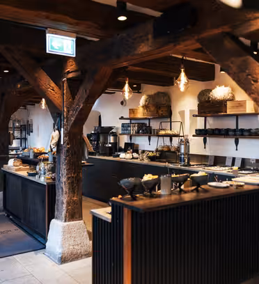 Spacious restaurant interior with rustic wooden beams and modern industrial lighting. Tables are set for dining, and buffets display a variety of foods. The atmosphere is warm, combining traditional and contemporary design elements.
