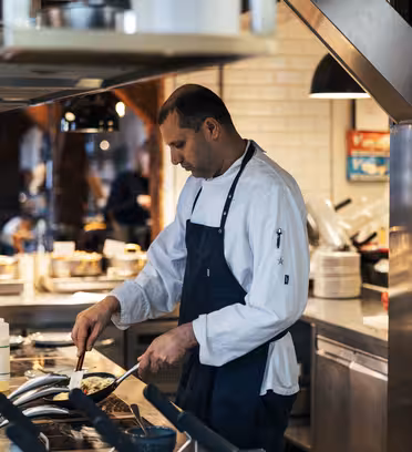 A chef in a white coat and dark apron is cooking on a stovetop in a modern restaurant kitchen. He is focused on preparing food in a pan, surrounded by kitchen equipment and ingredients, with shelves and lights visible in the background.