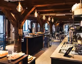 A spacious, warmly lit restaurant interior with rustic wooden beams and hanging lights. Buffet stations with various dishes line the sides, and tables are set for dining. Large windows offer natural light, adding to the inviting atmosphere.