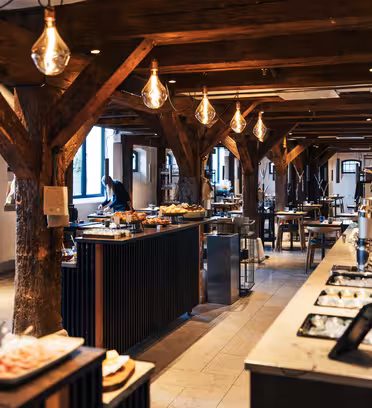 A spacious, warmly lit restaurant interior with rustic wooden beams and hanging lights. Buffet stations with various dishes line the sides, and tables are set for dining. Large windows offer natural light, adding to the inviting atmosphere.