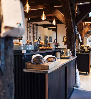 A rustic bakery interior with wooden beams and hanging lights. Freshly baked bread is displayed on wooden tables. The open kitchen area is visible in the background, with pots and pans. A cozy and inviting atmosphere.