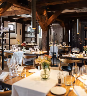 A cozy, elegant restaurant interior with wooden beams and white tablecloths. Tables are set with wine glasses, cutlery, and floral decorations. Warm lighting creates an inviting atmosphere. Empty chairs are neatly arranged around the tables.