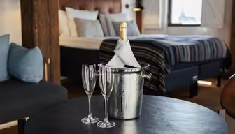 A cozy hotel room with a bed in the background. In the foreground, a bottle of champagne wrapped in a napkin is chilling in an ice bucket on a table, accompanied by two empty champagne flutes. A soft chair with cushions is to the left.