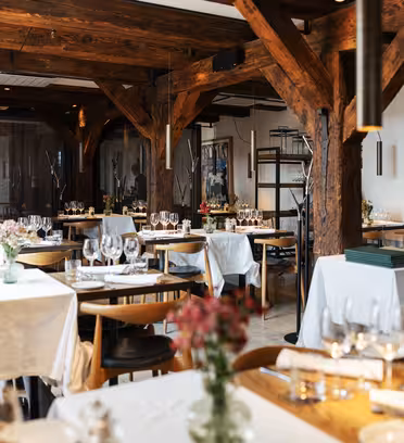 A cozy restaurant with wooden beams and pillars, featuring elegantly set tables with white tablecloths and floral arrangements. Soft lighting creates a warm ambiance, and delicate chandeliers are suspended from the ceiling.