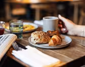 A hand holding a mug near a breakfast plate with a croissant, roll, and pastry. Nearby are glasses of juice and water, a newspaper, and silverware wrapped in a napkin on a wooden table. The background is softly blurred.