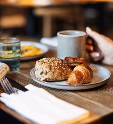 A hand holding a mug near a breakfast plate with a croissant, roll, and pastry. Nearby are glasses of juice and water, a newspaper, and silverware wrapped in a napkin on a wooden table. The background is softly blurred.