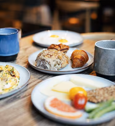 A breakfast spread on a wooden table, featuring a cheese omelette, plates of bread and croissants, sliced cheese with fruits and vegetables, and cups of coffee and juice.