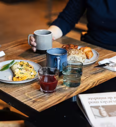 A person sits at a wooden table in a cafe with a mug, a plate of omelette and avocado, a plate with croissants and bread. There are glasses of juice and water, utensils wrapped in napkins, and a folded newspaper on the table.