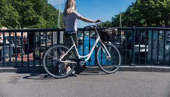 A person with long hair stands on a bridge beside a white bicycle. They are gazing at a canal lined with boats and trees under a clear blue sky. The scene is peaceful and sunlit.