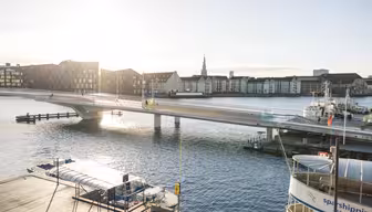 A modern pedestrian and cyclist bridge spans a calm, sunlit canal. The cityscape features historic buildings under a clear sky. Boats are moored on the water, and faint reflections shimmer on the canals surface.