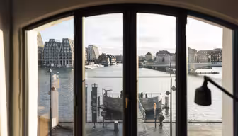 A view through an arched window shows a waterfront scene with modern and traditional buildings. A calm river reflects the cloudy sky, while a boardwalk full of people brings the tranquil cityscape to life.