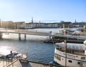 A scenic view of a harbor in Copenhagen, with a footbridge filled with people walking and cycling. Boats dock nearby and historic buildings line the water under a clear blue sky.
