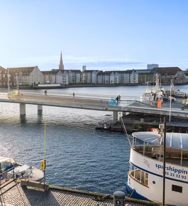 A scenic view of a harbor in Copenhagen, with a footbridge filled with people walking and cycling. Boats dock nearby and historic buildings line the water under a clear blue sky.