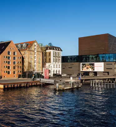 Waterfront scene with modern glass buildings and rustic brick structures along the quayside under a clear blue sky. Docked boats can be seen in the foreground, reflecting the calm water.