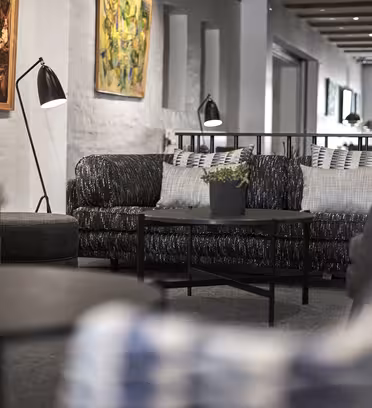 A stylish, modern living room features dark plush sofas, a black coffee table with a potted plant, and contemporary artwork on the walls. The room is well-lit with floor lamps, and large windows at the back allow natural light to brighten the space.