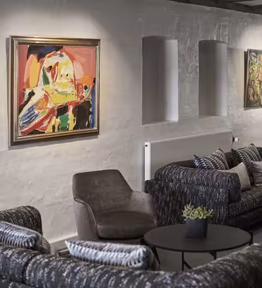 A cozy lounge with dark sofas and chairs arranged around round tables. The wall displays colorful abstract paintings, and soft lighting adds warmth. Small plants on tables enhance the inviting atmosphere.