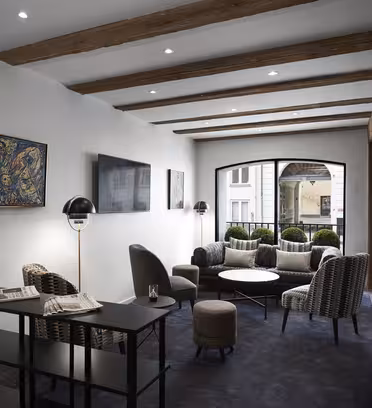 Modern lounge area with wooden beams and recessed lighting. Features cushioned chairs, a round coffee table, a floor lamp, and framed artwork on the walls. Black console table with newspapers is near large windows overlooking a street.