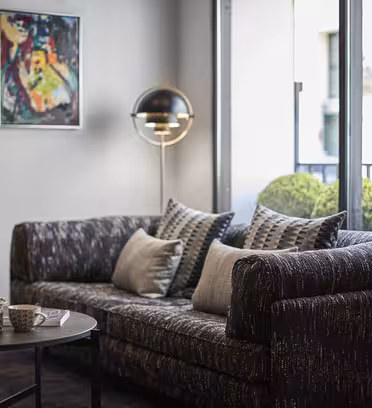 A modern living room with a dark patterned sofa adorned with pillows, a round coffee table with cups, and a spherical floor lamp. A colorful abstract painting hangs on the wall, and a large window reveals a patio with greenery outside.