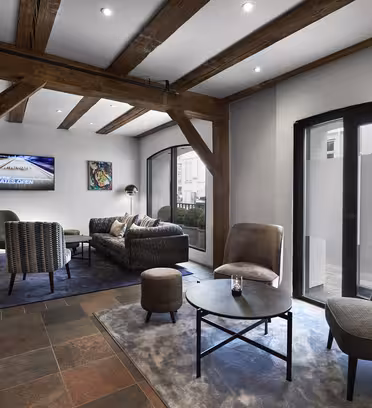 Modern lounge with exposed wooden beams, a mix of chairs and sofas, and a round coffee table on a patterned rug. A large TV is mounted on the wall, and artworks are displayed. Arched windows allow natural light to enter the space.