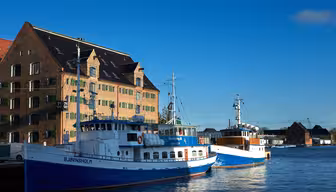 Two blue and white boats moor at a pier in a sunlit harbor. Behind them, a large brick building with green-framed windows rises against a clear blue sky. The water reflects the buildings and boats in this tranquil scene.