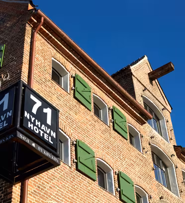 Brick building under a clear blue sky, featuring green shutters and a black sign with white text reading 71 Nyhavn Hotel. The buildings corner juts out, displaying the sign prominently.
