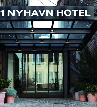 Entrance to 71 Nyhavn Hotel, featuring a glass canopy with recessed lighting. The entry is flanked by potted plants on either side, set against a backdrop of brick walls.
