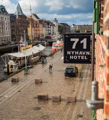 View of a cobblestone street along a canal in Copenhagen, with colorful buildings and boats lined up. A sign for 71 Nyhavn Hotel hangs on a building. Two people walk along the street under a partly cloudy sky.