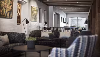 A cozy, modern lounge area with patterned sofas and round black tables. Artwork adorns the white brick walls. Ceiling beams add charm, and a large window at the back lets in natural light. Potted plants decorate the tables.