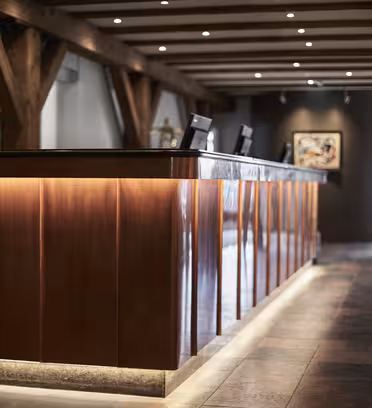 A sleek, modern hotel reception area with a wooden front desk and soft, warm lighting. Two computer monitors are on the desk. The walls are decorated with contemporary artwork, and wooden beams are visible on the ceiling.