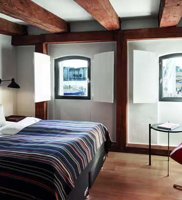 Cozy bedroom with a double bed featuring a striped bedspread and two pillows. Exposed wooden beams on the ceiling. Two small arched windows provide a view of a waterfront and buildings. A desk with two chairs is placed near the windows.