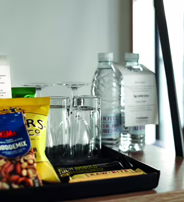 A tray on a table holds a red wine bottle, two water bottles, four empty glasses, and various snacks including a bag of mixed nuts and chocolate bars. A note is placed next to the wine.