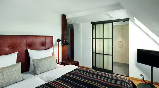 A modern hotel room features a large bed with a brown headboard and striped bedding. Two pillows rest on the bed. A black floor lamp stands by a wooden beam. A flat-screen TV is opposite the bed, near a sliding glass door leading to a bathroom.