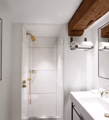 A modern bathroom with a white interior, wood beam ceiling, and gold fixtures. It features a glass-enclosed shower, large mirrored wall with two towel racks, and a white sink with a sleek counter.