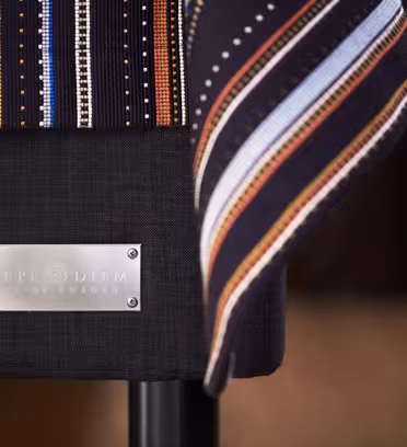Close-up of a fabric with vertical stripes in various colors draping over a piece of furniture. A metal plaque on the furniture reads Carpe Diem Beds of Sweden. The background is out of focus.