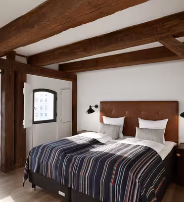 A cozy bedroom with a large bed covered in a striped blanket. Wooden beams adorn the ceiling, and two wall-mounted lamps flank the bed. Light wood flooring complements the rustic decor, with large windows providing natural light.