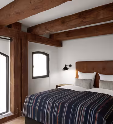 A cozy bedroom with a double bed featuring a striped duvet and two pillows. The room has wooden beams on the ceiling, a large arched window, and two wall-mounted lamps. The decor is minimalist with a rustic touch.