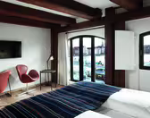 Hotel room with large wooden beams, a bed with a striped blanket, two brown chairs, a small desk with a lamp, and a wall-mounted TV. Large windows provide a view of the cityscape and waterfront.