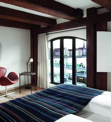 Hotel room with large wooden beams, a bed with a striped blanket, two brown chairs, a small desk with a lamp, and a wall-mounted TV. Large windows provide a view of the cityscape and waterfront.