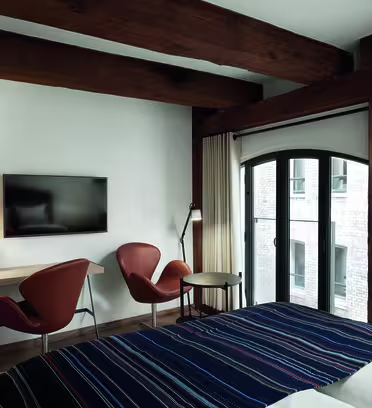 Hotel room with a bed featuring a striped blanket, two orange chairs, a small round table, a wall-mounted TV, and wooden beams on the ceiling. The room has a large window with curtains partially drawn.