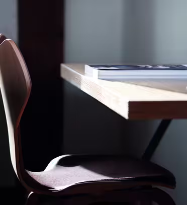 A wooden chair with a curved back sits next to a wooden table in soft lighting. A closed magazine lies on the tables surface. The setting appears minimalistic and cozy.