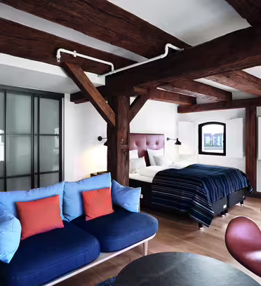 A modern bedroom with exposed wooden beams features a double bed, a blue-striped comforter, a blue sofa with orange cushions, a brown chair, and a small round table. Large windows and a sliding glass door enhance the space.