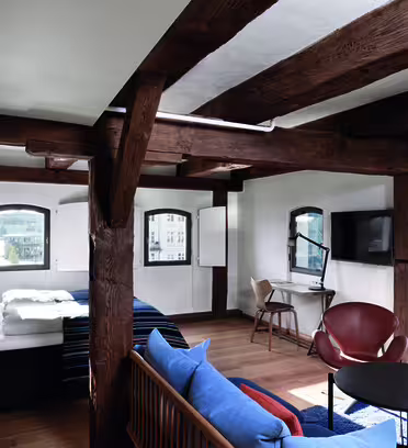 A rustic loft with wooden beams features a bed, a cozy seating area with a couch and chairs, a round coffee table, and a desk with a TV. Large windows bring in ample natural light and offer a view of the city outside.