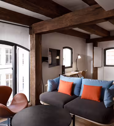 A cozy room with exposed wooden beams features a modern sofa with blue and orange cushions, a round black table, an armchair, and a work desk next to a window. Large windows offer a view of an outside building.