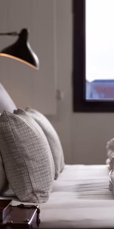 A cozy bedroom with a neatly made bed, featuring white linens and gray cushions. Two modern black lamps are mounted on the padded headboard. A window in the background shows a blurred cityscape with a prominent spire.